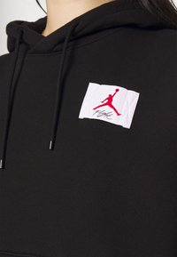 Sudadera negra con un parche acolchado blanco que presenta un logo Jumpman rojo y el texto "Flight". Los cordones tienen puntas de metal. Textura suave.