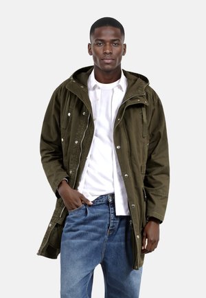 Parka verde oliva con capucha, cremallera frontal, automáticos y bolsillos laterales, llevada sobre una camisa blanca y jeans azules.