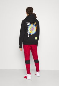 Svart huva sweatshirt med en färgglad basketgrafik och texten "VEM?" tillsammans med röda byxor med svarta ränder och vita skor.