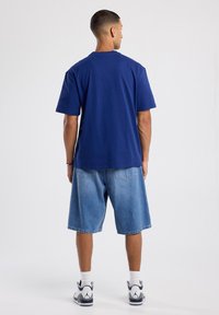 T-paita tummansinisestä puuvillasta, lyhyt hiha ja pyöreä kaula-aukko. Yhdistetty vaaleansiinisiin farkkushortseihin ja valkoisiin lenkkareihin.