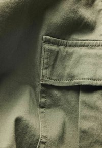 Gros plan d'une poche en tissu vert avec coutures visibles et un rabat, partie d'un vêtement en coton texturé.