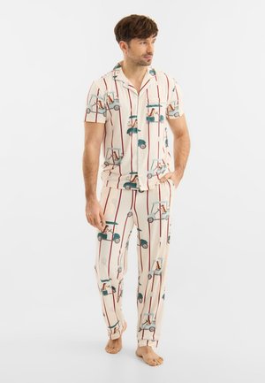 Mand iført cremefarvet pyjamas med røde striber og golfvognsmønster, stående barfodet mod en ensfarvet baggrund.