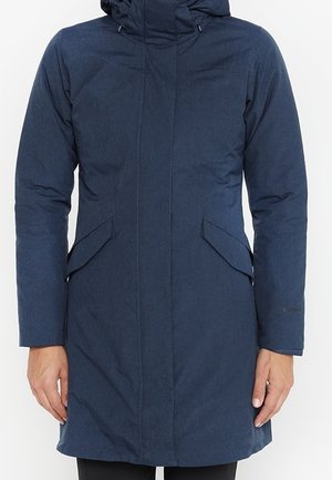 Marineblauwe lange winterjas met capuchon, voorflapzakken en verborgen rits, gedragen door een persoon met ontspannen handen langs het lichaam.