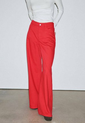 Persona que lleva pantalones rojos brillantes de pierna ancha y una camiseta blanca ajustada de manga larga, de pie con las manos detrás de la espalda contra un fondo liso.