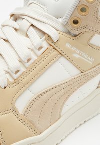 Béžové a bílé sneakersy s texturovaným semišem a koženými materiály, mají kulatou špičku, bílé šněrování a detail s logem "SLIPSTREAM".