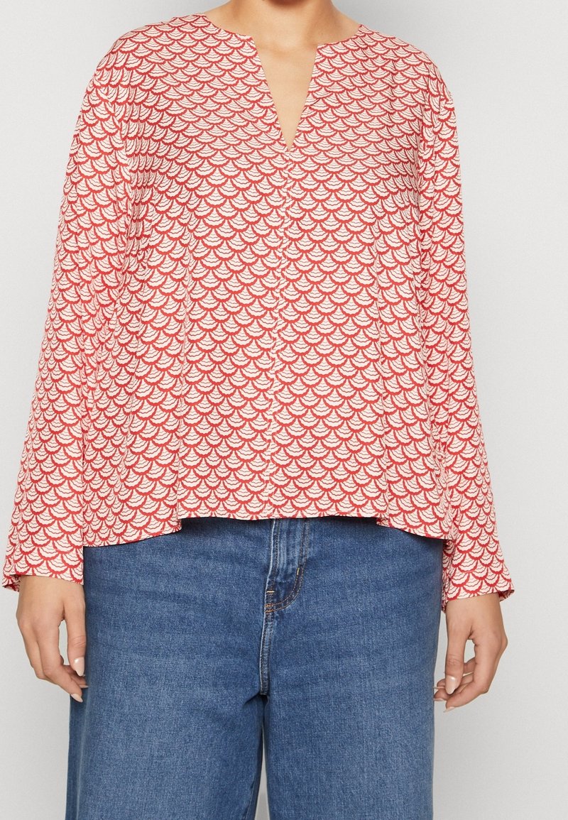 Femme portant une blouse à manches longues rouge et blanche à motif avec un décolleté en V et un jean en denim bleu, se tenant devant un fond uni.