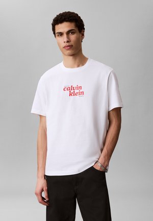 Giovane uomo in piedi con una mano in tasca, indossa una t-shirt bianca Calvin Klein con logo rosso e pantaloni neri su sfondo grigio uniforme.