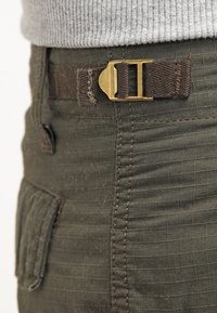 Olivgrüne Hose aus strukturiertem Stoff, mit einer flachen Tasche und einem braunen Textilgürtel mit einer Metallschnalle und verstärkten Nähten.