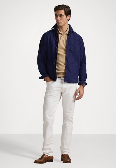 Chaqueta ligera navy con cierre de cremallera, polo beige debajo, pantalones blancos y zapatos de cuero marrón. Diseño minimalista, forma ajustada.