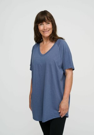 Glunderende vrouw met bruin haar die een loszittend blauw topje met korte mouwen en een zwarte broek draagt, staat tegen een effen lichte achtergrond.