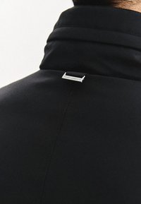 Czarna kurtka z wysokim kołnierzem, gładką fakturą i srebrną metką marki z tyłu. Projekt jest elegancki i nowoczesny.