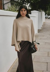 Un poncho de punto beige cae sobre una falda maxi de satén marrón oscuro. El conjunto incluye un bolso negro con un detalle de hebilla plateada.