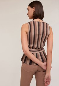 Top fără mâneci, cu dungi verticale maro, de culoare bej, cu o fustă evazată și talie strânsă. Modelul poartă pantaloni maro, cu mâinile la spate.