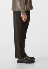 Pantalones de pierna ancha de color marrón oscuro, hechos de una tela suave, combinados con zapatos negros voluminosos que presentan un detalle de hebilla y suelas texturizadas.