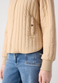Veste matelassée beige avec zip et détail de poche à pression, portée avec un jean denim bleu, visible du milieu du torse au poignet.