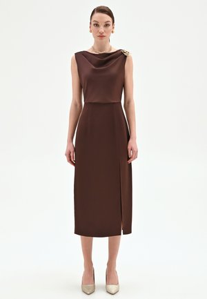 Donna che indossa un abito midi senza maniche marrone con scollo a collo a drappeggio, spilla dorata sulla spalla, collana d'oro, orecchini e tacchi dorati appuntiti.