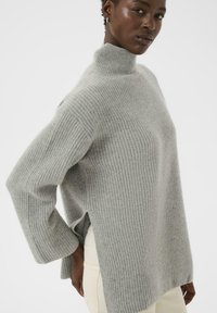 Woman wearing a light gray ribbed high-neck sweater with side slits, paired with light-colored pants, against a plain white background.