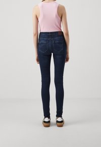 Jeans ajustados de color azul oscuro con bolsillos traseros y un parche de cuero en la cintura, usados con una camiseta sin mangas de color rosa claro y zapatos blancos.