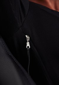 Κοντινή λήψη ενός ασημένιου φερμουάρ σε μαύρο ριμπ ύφασμα με μαύρο κορδόνι έλξης, εν μέρει καλυμμένο από μαύρο υλικό με υφή.