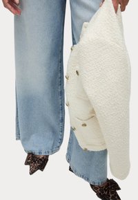 Chaqueta blanca texturizada sostenida en una mano, con botones dorados. Un par de jeans de pierna ancha en azul claro son visibles, junto con una bufanda de estampado animal oscuro.