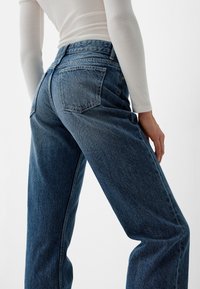 Persona con pantalones vaqueros de cintura alta, de corte holgado, y una camiseta blanca de manga larga ajustada, posando con una mano cerca del muslo.