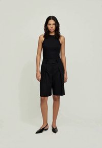 Top justo sem mangas preto combinado com calções altos pretos, apresentando detalhes em pregas e sapatos pretos de bico fino.