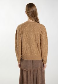 Jasnobrązowy sweter z dzianiny z teksturowanym wzorem w diamenty i ściągaczem na dole, noszony na wzorzystej brązowej sukience z falbankami.