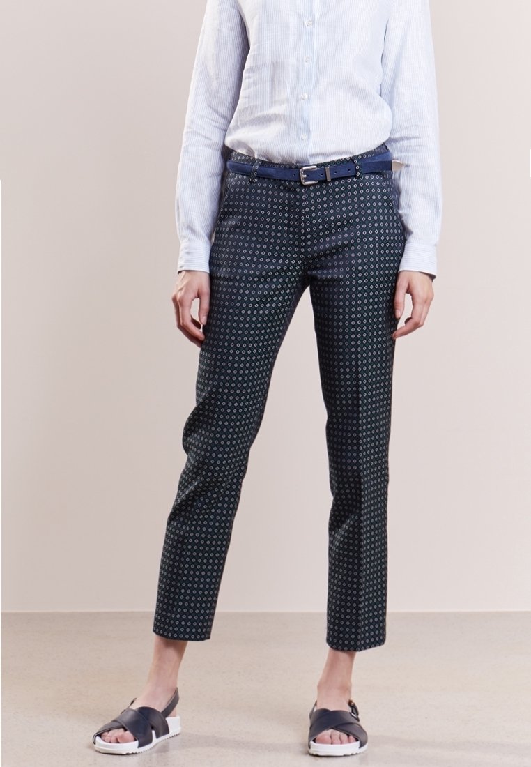 Femme portant un pantalon slim foncé à motif, chemise bleu clair à manches longues rentrée, ceinture bleu marine et sandales noires à bouts ouverts.