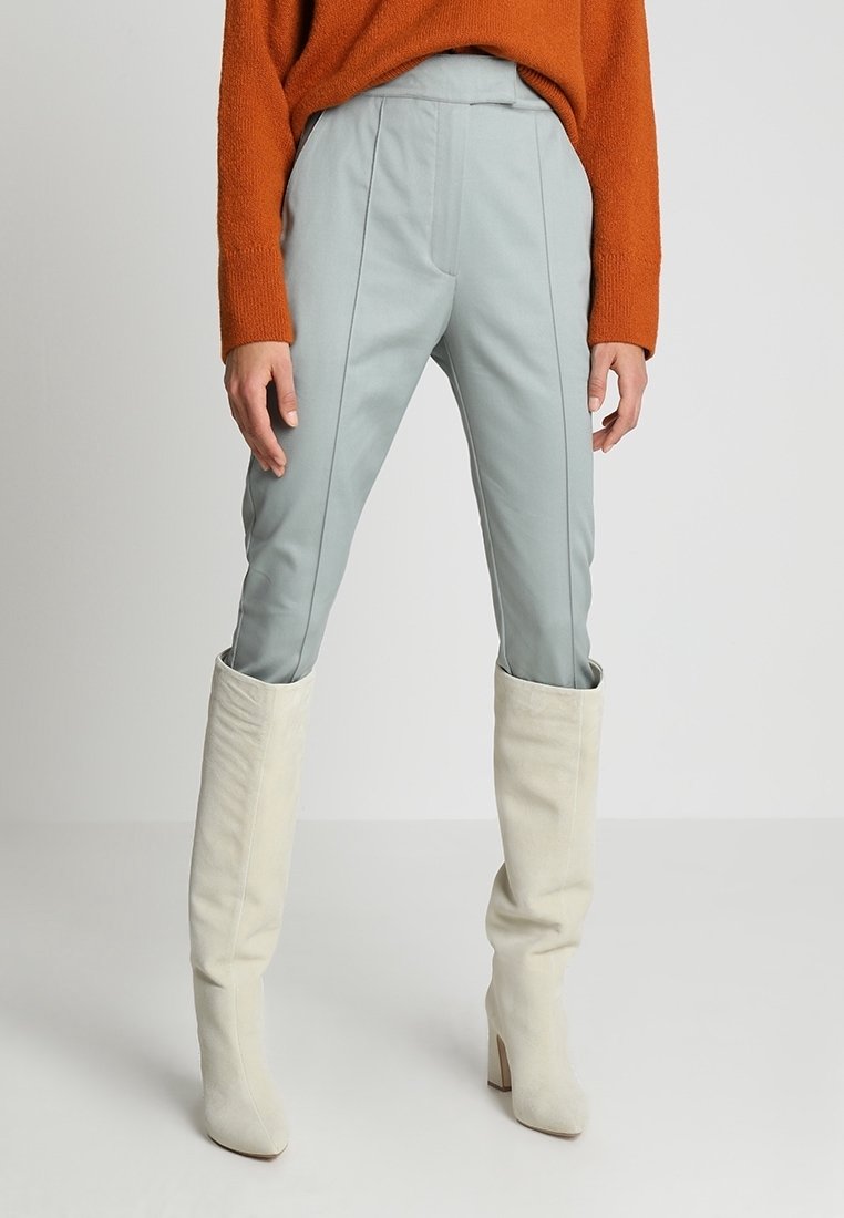 Světlé modré kalhoty na míru s bočními švy, spárované s bílými vysokými kozačkami. Oblečení doplňuje živý oranžový strukturovaný svetr.