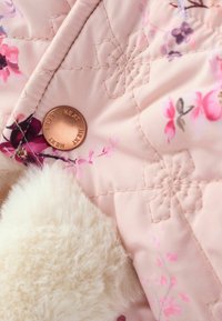 Gesteppte rosa Jacke mit floralem Muster, ausgestattet mit einem Metallknopf und weichem weißen Kunstfellbesatz. Sichtbare Nähte und Details betonen die Textur.