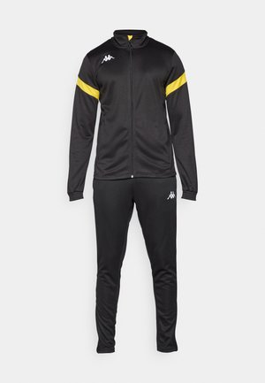 Svart atletisk treningsdrakt med en jakke med glidelås og høy krage, gule striper på skuldrene, og matchende bukser. Glatt tekstur.