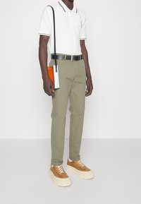 Biała koszulka polo z czarnym obszyciem, oliwkowozielone spodnie, beżowe buty sportowe z białymi podeszwami oraz pomarańczowo-biała torba na ramię.