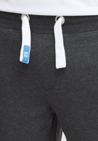 Pantalon de survêtement gris avec un cordon blanc, portant une étiquette bleue portant la mention "SLD". Texture douce avec une taille élastique et une coupe classique.