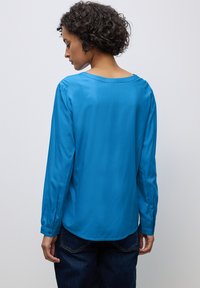 Blauwe blouse met lange mouwen, een brede halslijn en een gebogen zoom. Gladde stof met een beetje glans. De manchetten hebben een knoopdetail. G gedragen met donkere spijkerbroeken.