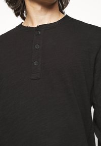 Maglietta henley nera a maniche lunghe con una finitura testurizzata, caratterizzata da un collo rotondo, bordi sfilacciati e tre bottoni neri al colletto.