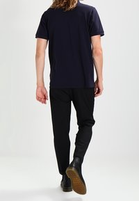 Laivastonsininen lyhythihainen t-paita, mustat slim fit -housut ja mustat kengät, joissa on teksturoitu pohja. Näkyvissä hiuksia ja neutraali tausta.