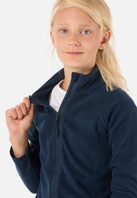 Laivaston fleece-takki, jossa on korkea kaulus ja täysimittainen vetoketju, ykkäisenä rintalogo ja joustavat hihansuut. Sujuva pinta, kevyt muotoilu.