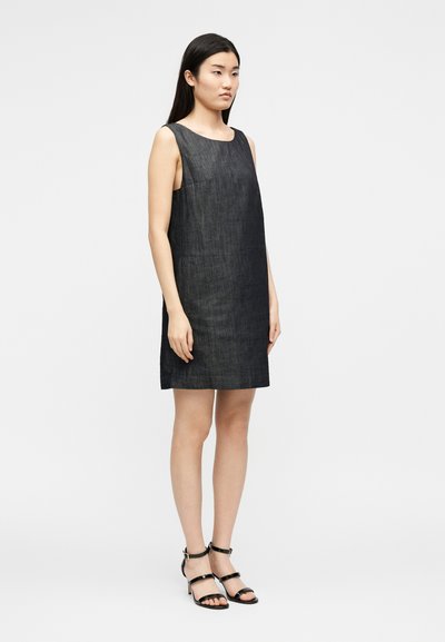 Γυναίκα με μακριά σκουρόχρωμα μαλλιά που φοράει αμάνικο σκουρογκρι φόρεμα έως το γόνατο και μαύρα πέδιλα με λουράκια, στέκεται μπροστά σε απλό ανοιχτό φόντο.