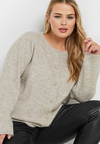 Beige stickad cardigan med rund halsringning, fem neutrala knappar och ribbade ärmslut. Materialet har en mjuk, strukturerad yta.