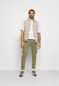 Beige Steppweste über einem weißen gestreiften Hemd, olivgrünen Cargo-Hosen und grauen Sneakers. Das Model steht vor einem schlichten weißen Hintergrund.