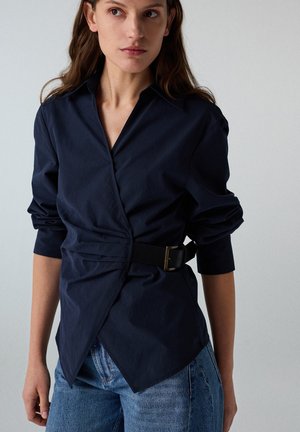 Mujer que lleva una camisa cruzada azul marino oscuro con un cinturón negro y mangas arremangadas, combinada con vaqueros azul claro, de pie frente a un fondo liso.