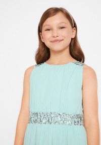 Un vestito senza maniche di colore verde menta con tessuto plissé, vita decorata con paillettes argentate e accenti decorativi sulle spalle.