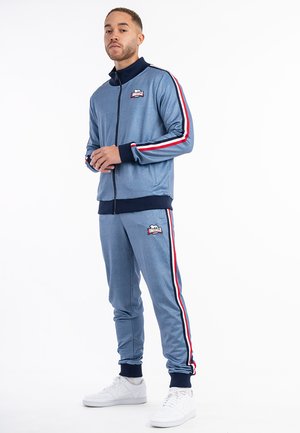 Tracksuit i lyseblåt stof med røde og hvide sidestropper; har en lynlåsjakke og tætsiddende bukser med logo-detaljer og ribbede manchetter.