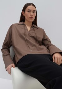 Bruine oversized blouse met een V-hals en trekkoord, met lange mouwen en een ontspannen pasvorm, gecombineerd met zwarte broeken.
