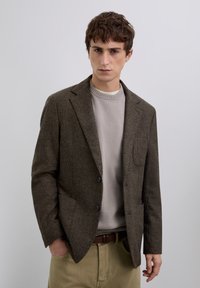 Hombre vestido con un blazer de cuadros marrón sobre un suéter beige y pantalones caqui, de pie con una mano en el bolsillo contra un fondo liso claro.