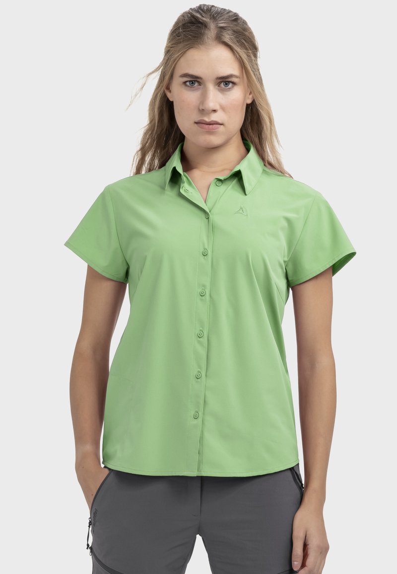 Frau mit langen blonden Haaren, die ein hellgrünes, kurzärmeliges Hemd mit Knöpfen und dunkelgraue Hosen trägt, steht mit den Händen in den Taschen.