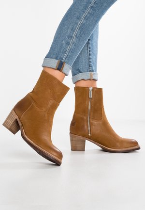 Bottes de cheville en cuir brun avec une finition lisse, tige de hauteur intermédiaire, fermeture éclair sur le côté et un talon empilé, positionnées sur un fond blanc.