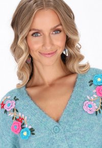 Svetlo modrý pletený cardigan s hlbokým V-výstrihom, zdobený farebnou kvetinovou výšivkou a jedným gombíkom na zapínanie. Viditeľná je jemná textúra.