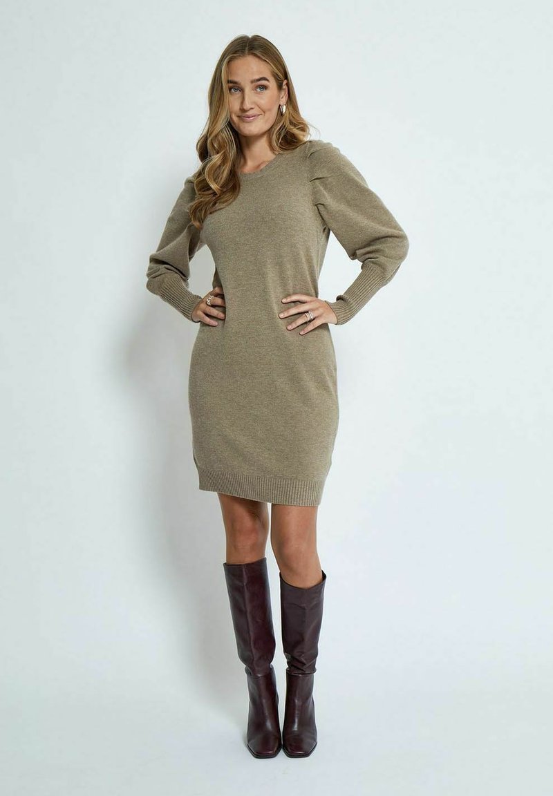Knit sweater dress in olive green, featuring a V-neck, puffed sleeves, ribbed cuffs, and hem. Paired with knee-high burgundy boots.