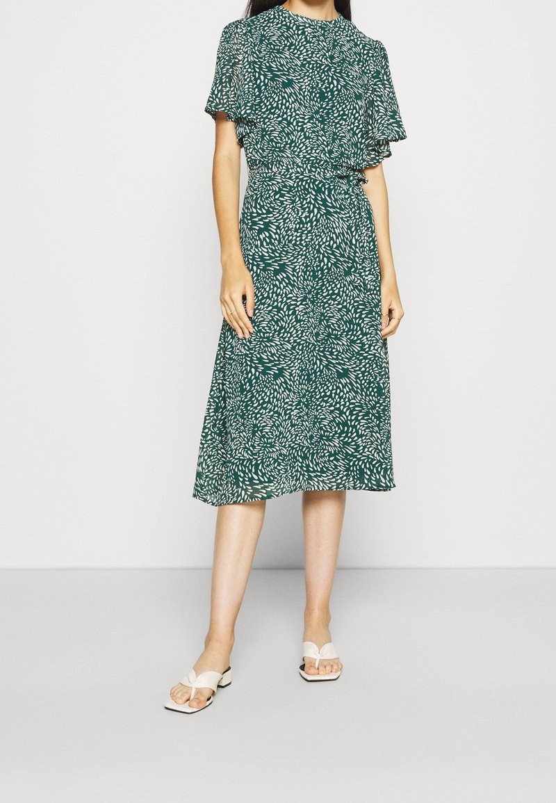 Grön klänning med vitt lövmönster, korta sleefar, åtstramad midja och svävande midilängd. Matchad med vita sandaler.